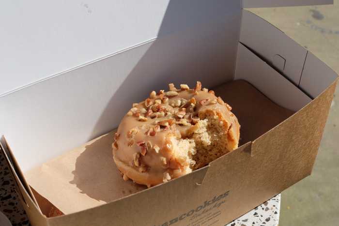 A maple pecan donut in a box with one bite taken out of it