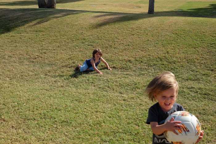 One kid laughing and holding the soccer ball while the other kid is on their knees getting up from the grass