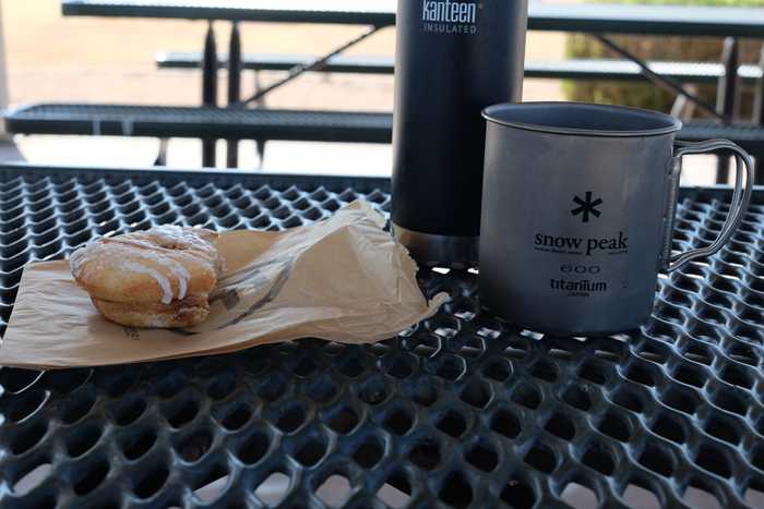 A close up of a donut, titanium mug, and insulated kleen kanteen sitting on a picnic table