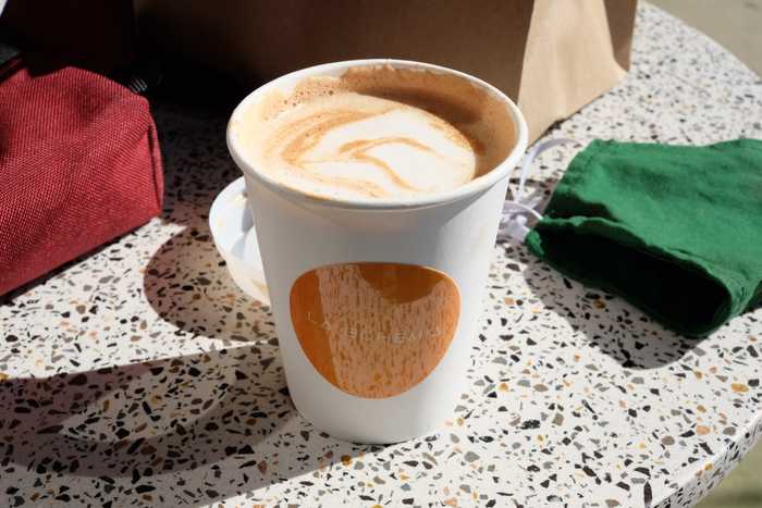 A mocha in a paper cup with an orange LA BOHEMIA sticker on it sitting on a table. In the background is a mask, hip pack, and paper bag.