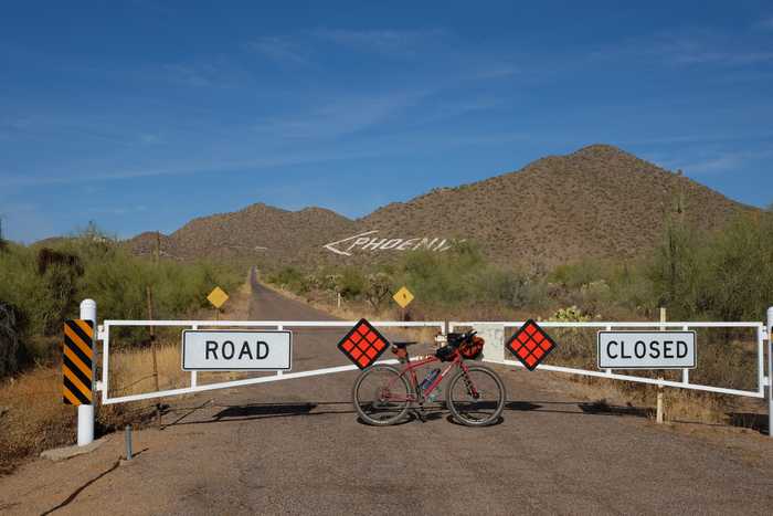 A red Surly Bridge Club leaning against a gate with a sign saying "Road Closed" and rocks painted to form an arrow and the word PHOENIX in the side of a mountain in the background