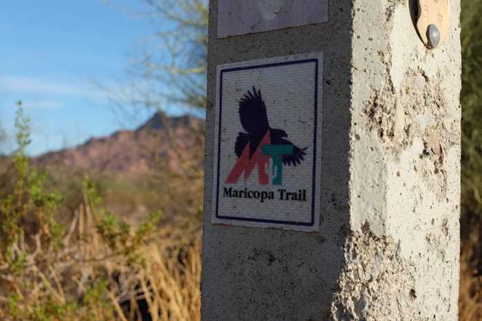 A close up of a Maricopa Trail sign along the canal with a blurry Red Mountain in the background