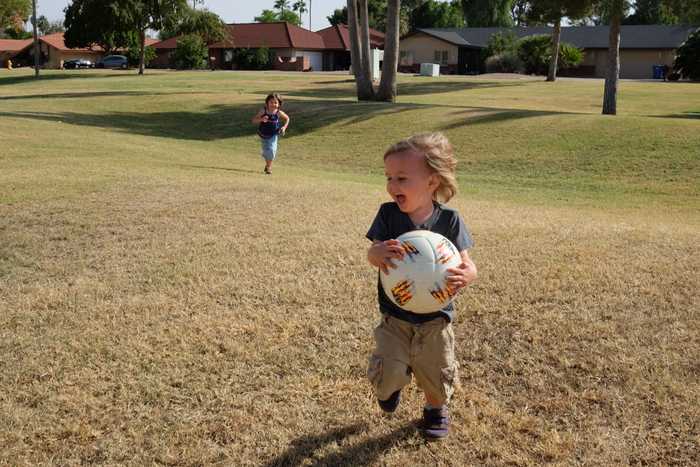 One kid laughing and running while holding the soccer ball as the other kid runs toward them